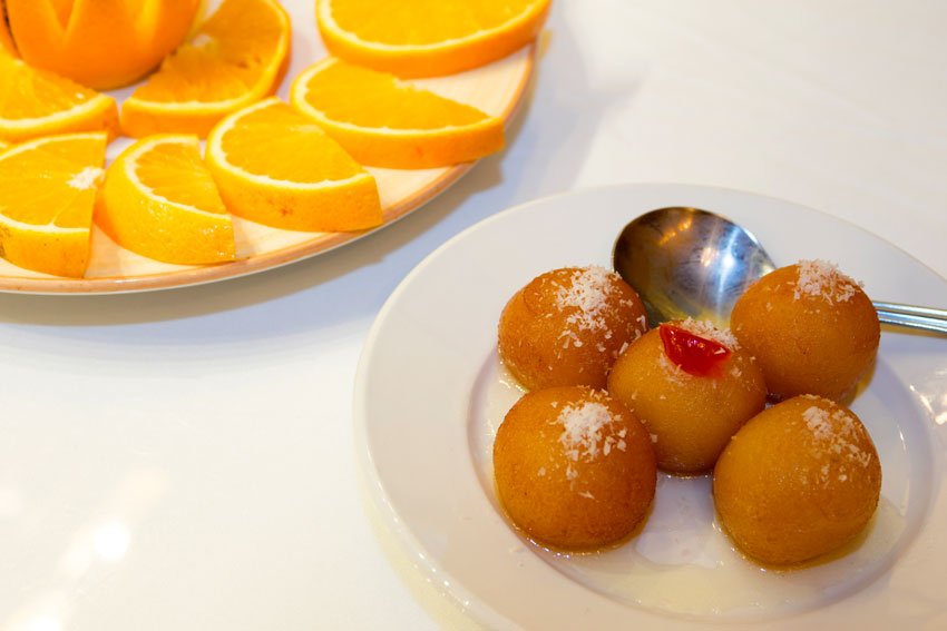 Taj of Marin Holiday Schedule - Desserts - Partial view of a plate of sliced oranges and a small plate of Gulab Jamun with a spoon. 